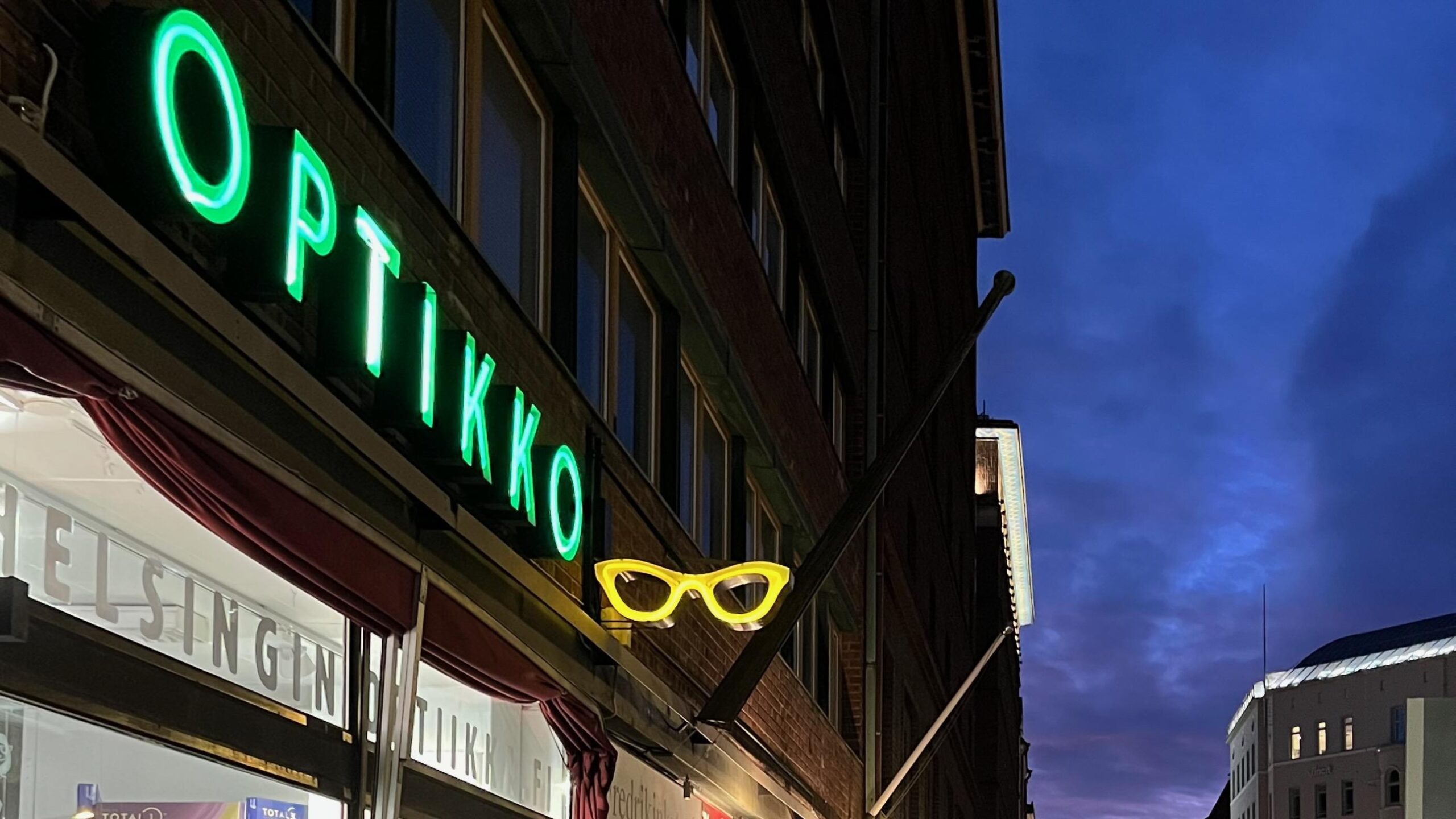 Helsingin Optiikka, the shop seen from the outside. The name of the shop is written with illuminated green letters.