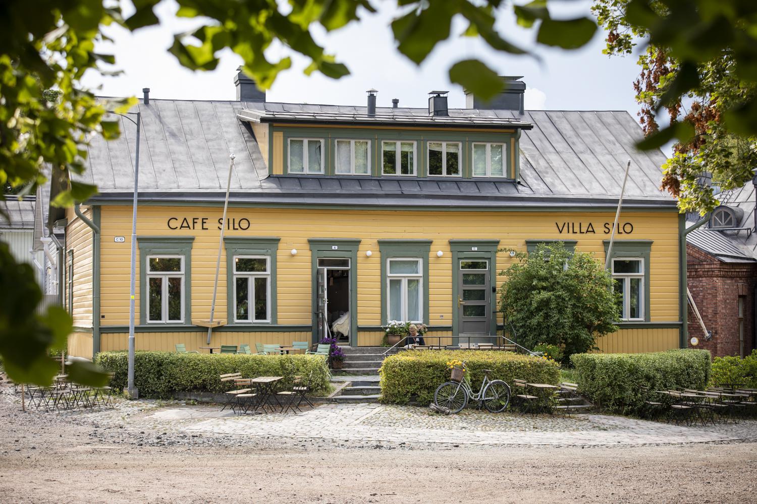 Puiden lehtien takana näkyy keltainen puurakennus, jonka edessä on vihreät puskat.