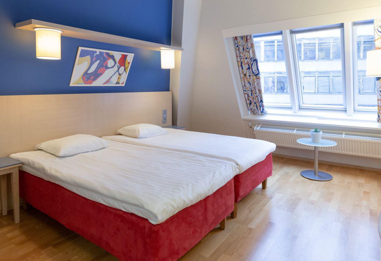 Scandic Hakaniemen hotellihuone. Sänky jossa punaista ja valkoista väriä, takana sininen seinä ja vieressä ikkunat.