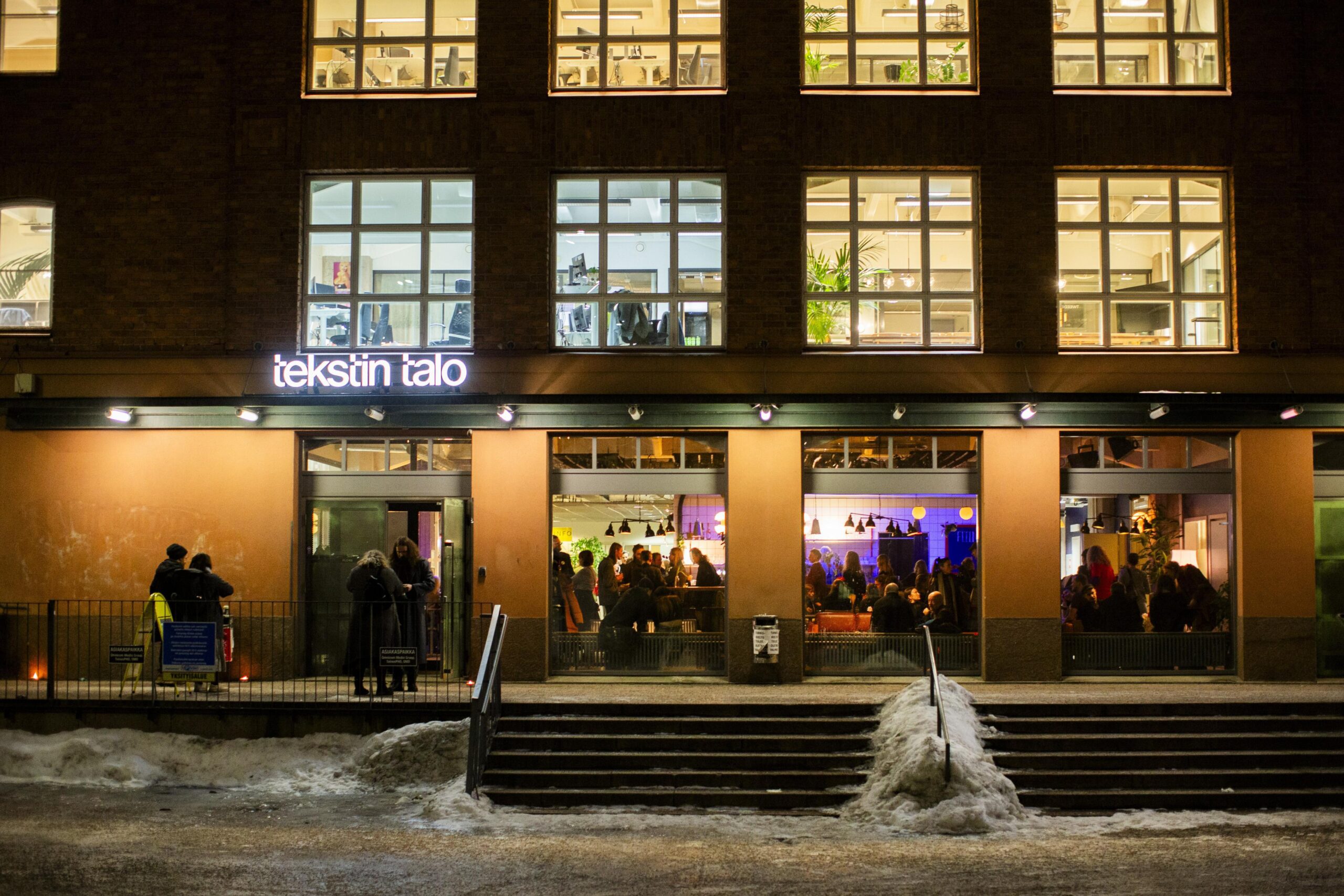 Kuvassa Tekstin talon julkisivu. Ikkunasta näkyy ihmisiä sisällä baarissa. Oven yllä hohtaa Tekstin talon nimi valokirjaimin.