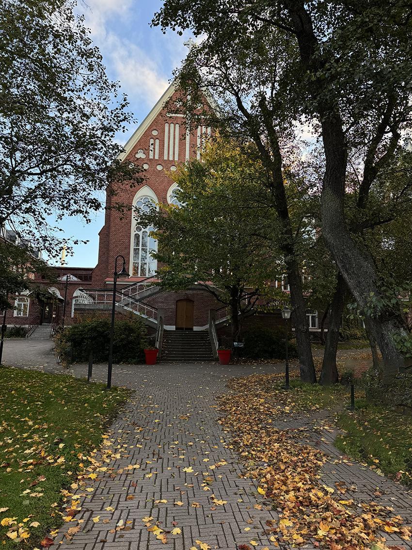 A red-brick building behind green leaves. Yellow leaves on the ground in the foreground.