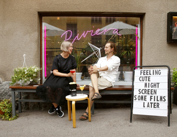 Couple enjoying drinks and popcorn outside Riviera cinema, with "Riviera" neon sign.