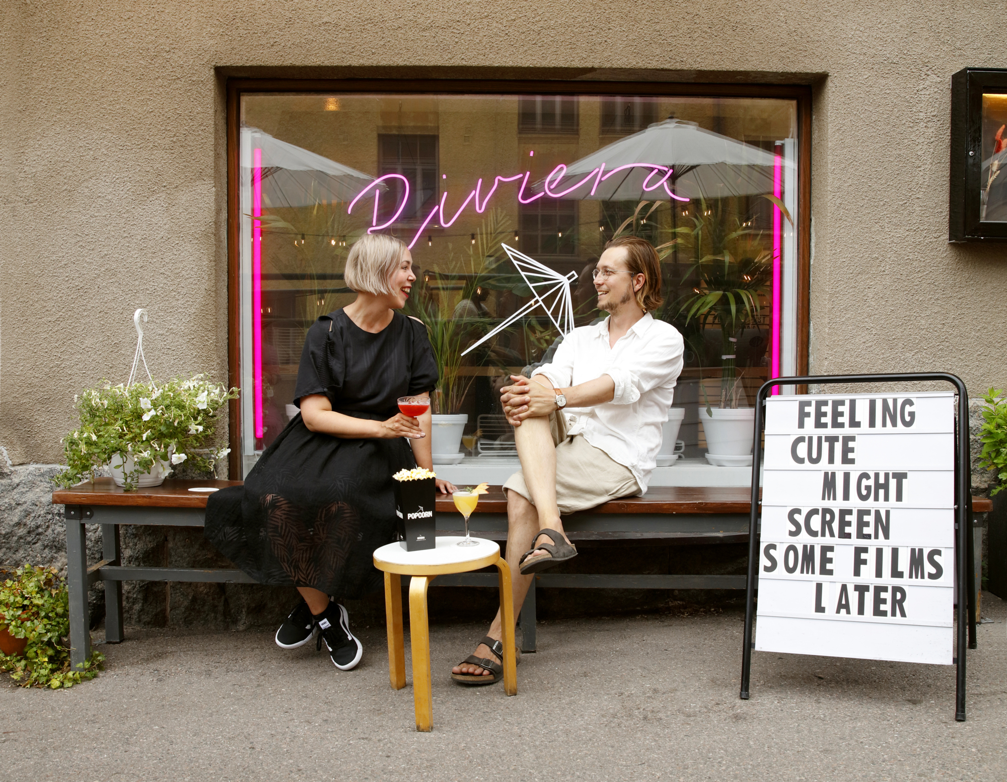Couple enjoying drinks and popcorn outside Riviera cinema, with "Riviera" neon sign.