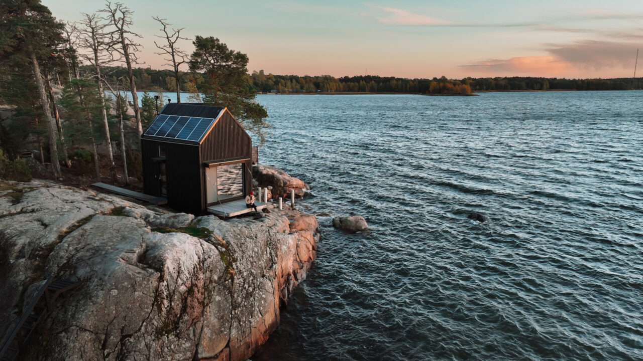 Modern black cabin with solar panels on rocky cliff overlooking a lake at sunset.
