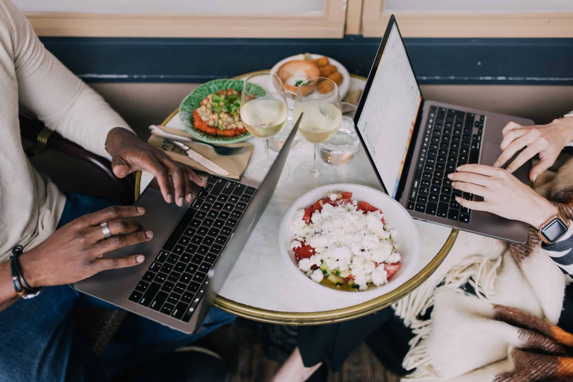 Two people working on laptops at a table with food and drinks.