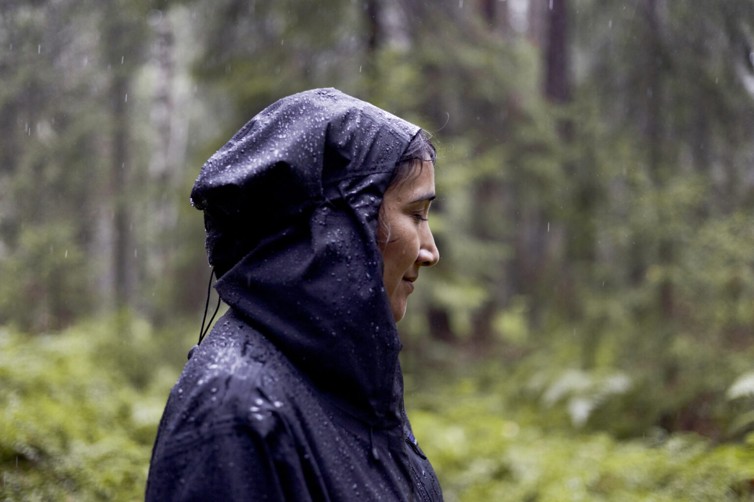 Woman in a black hooded jacket with raindrops in a green forest.