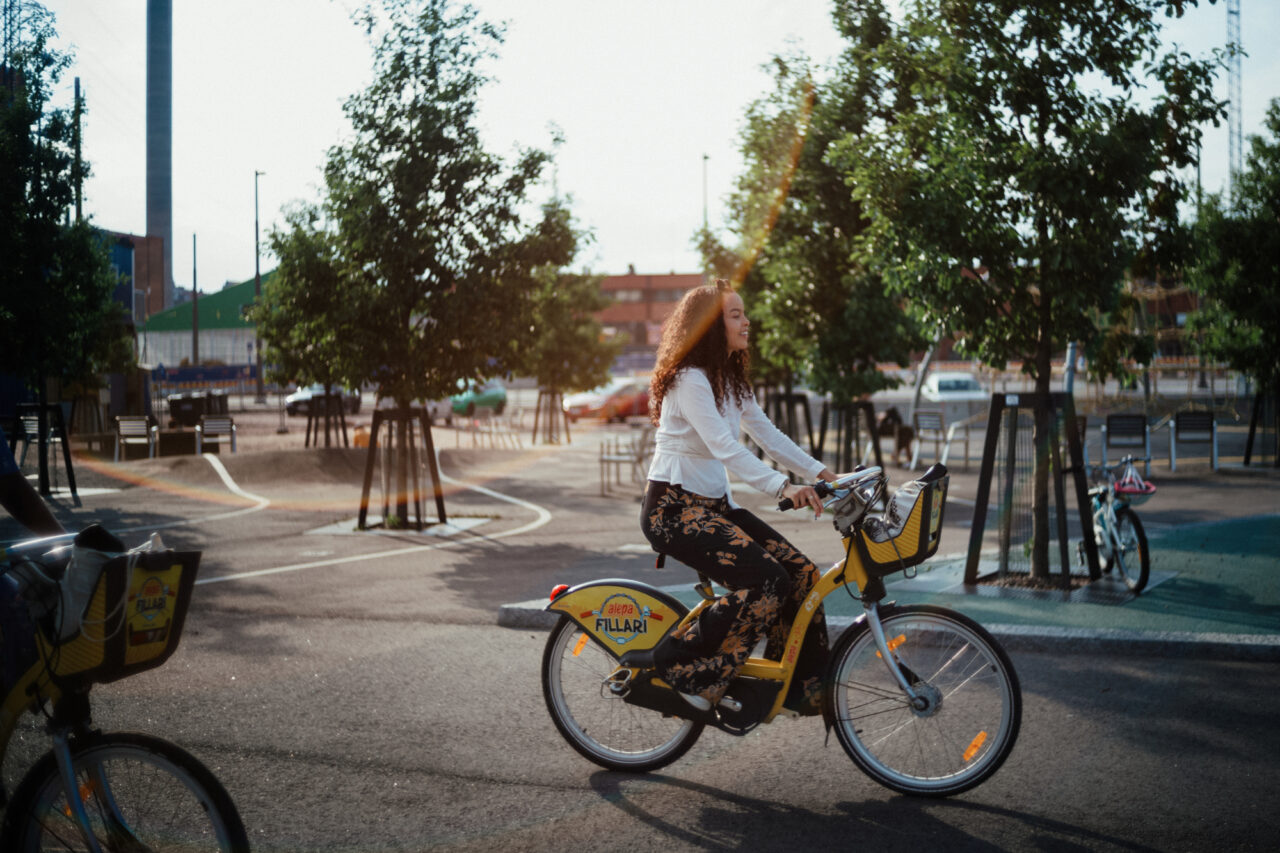 Woman riding a yellow shared bike with "Fillari" logo in a park.
