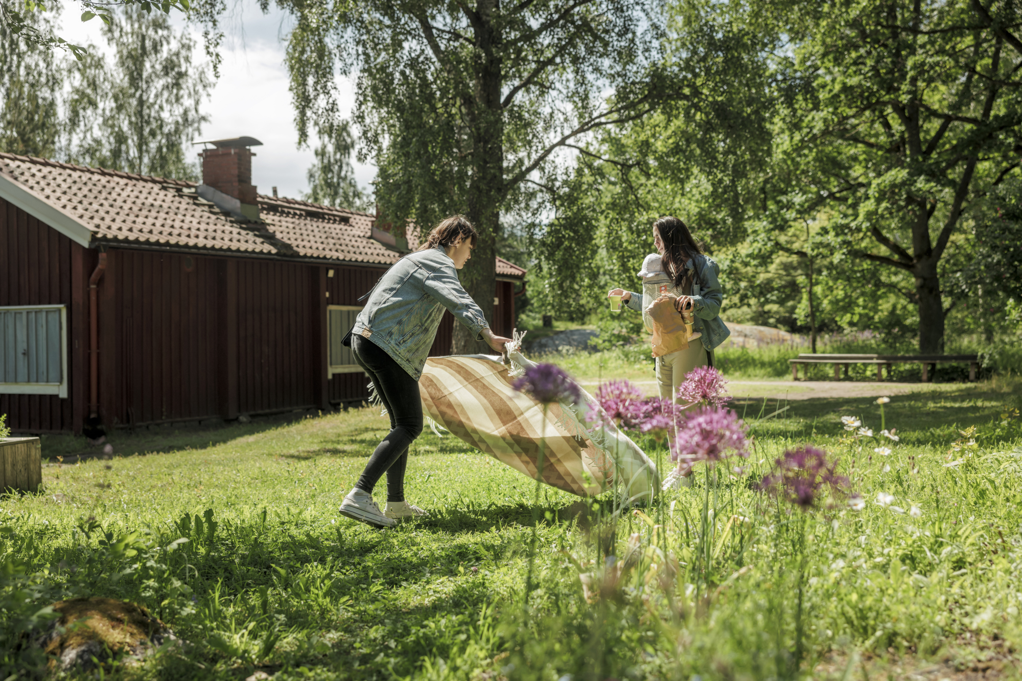 Two women setting up a picnic blanket in a sunny garden with a red cabin.