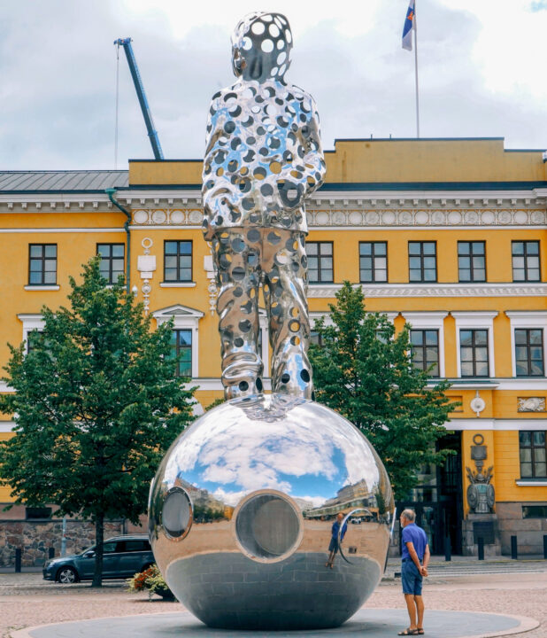 Giant mirrored sculpture of a person on a sphere in front of a yellow building