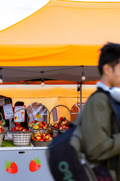 Fresh apples and strawberries at a vibrant outdoor market stall under an orange tent.