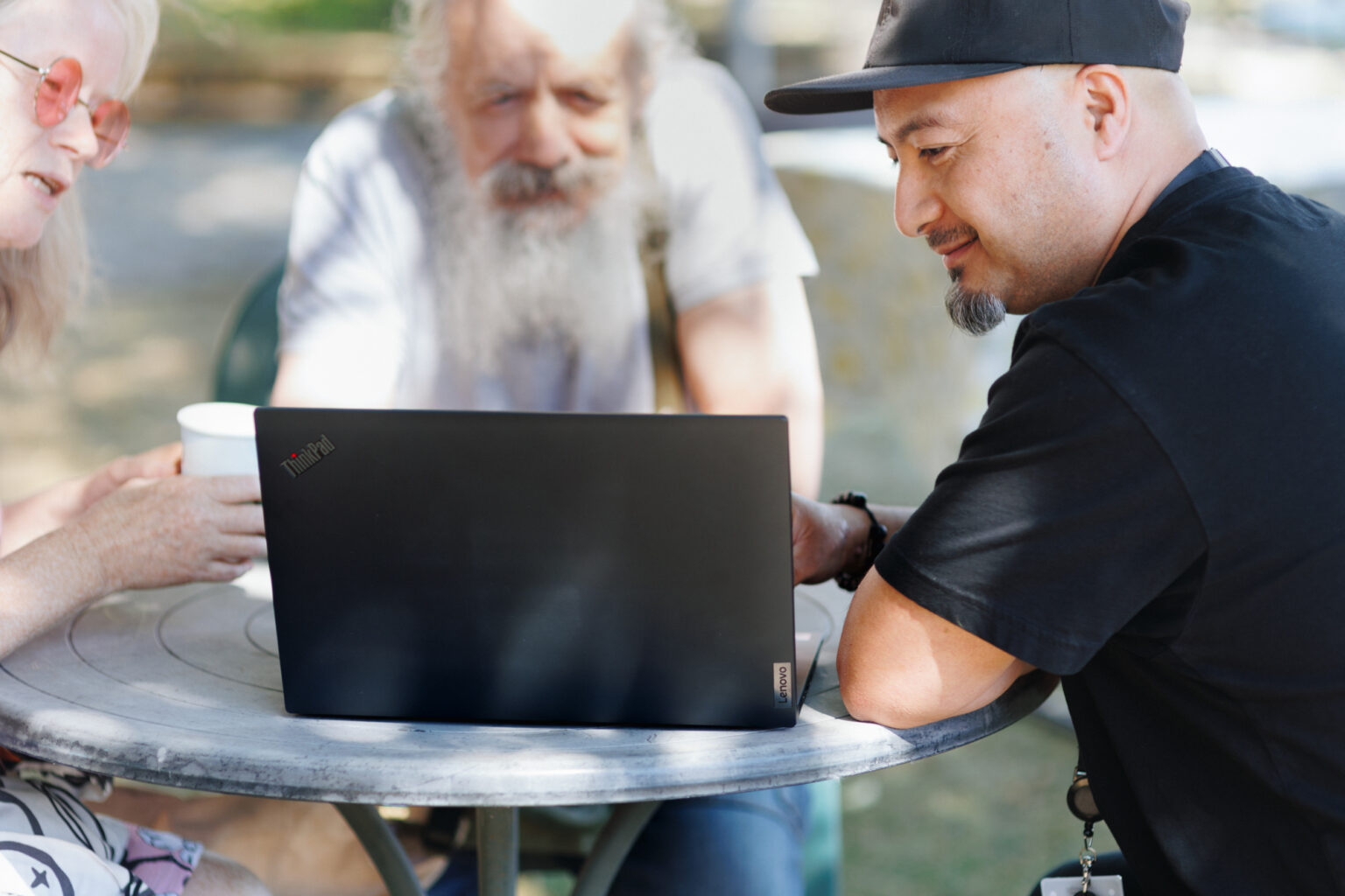 Man in black cap shows laptop screen to two people at outdoor table