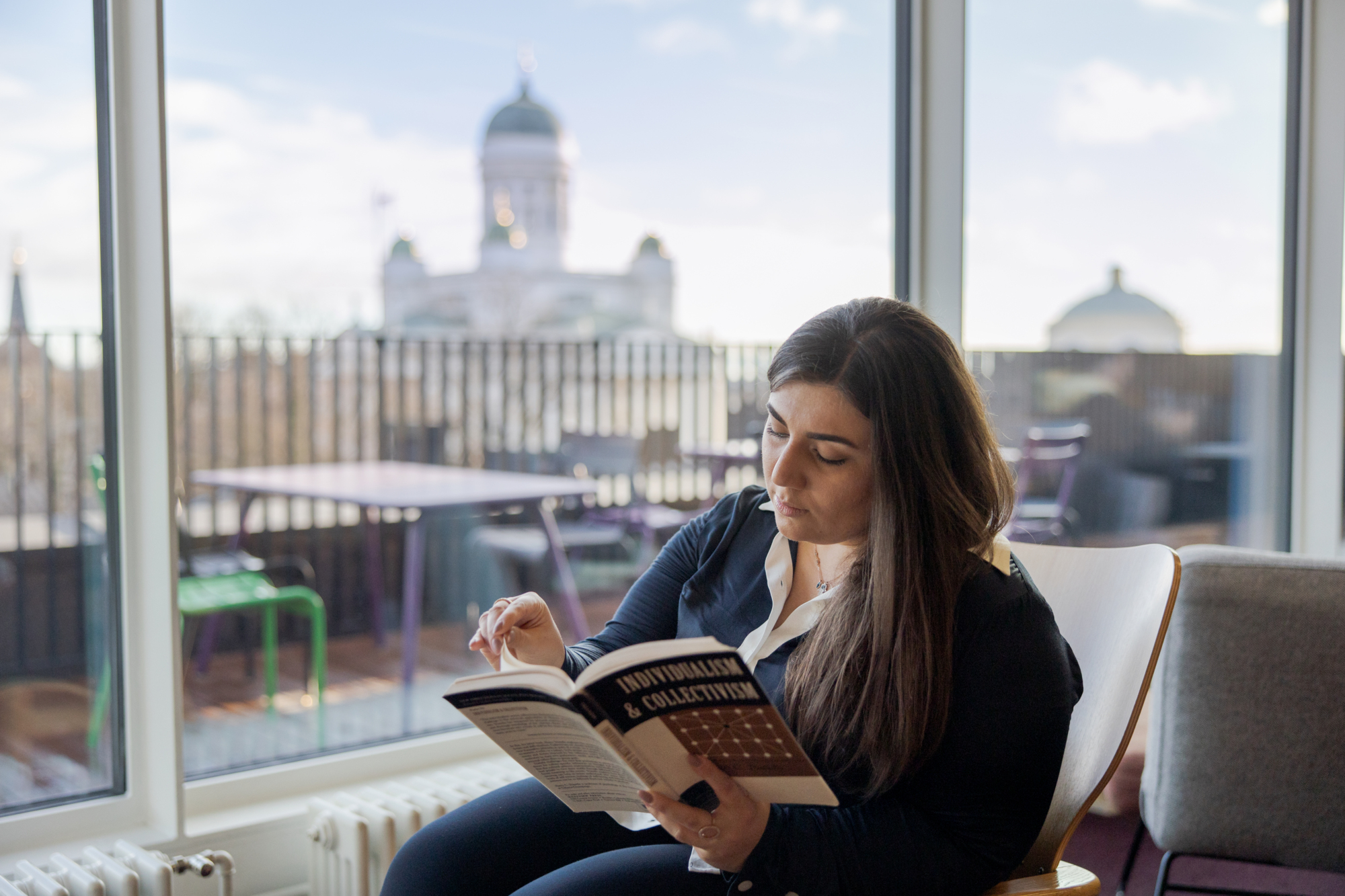 Woman reading "Individualism & Collectivism" by large window with Helsinki Cathedral view