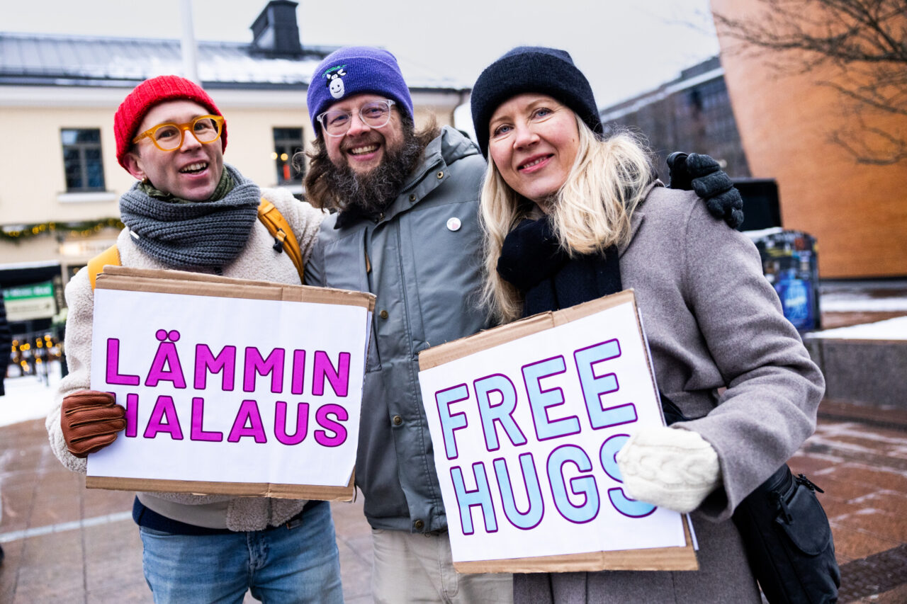 A group of three friends holding signs about hugs in Helsinki.