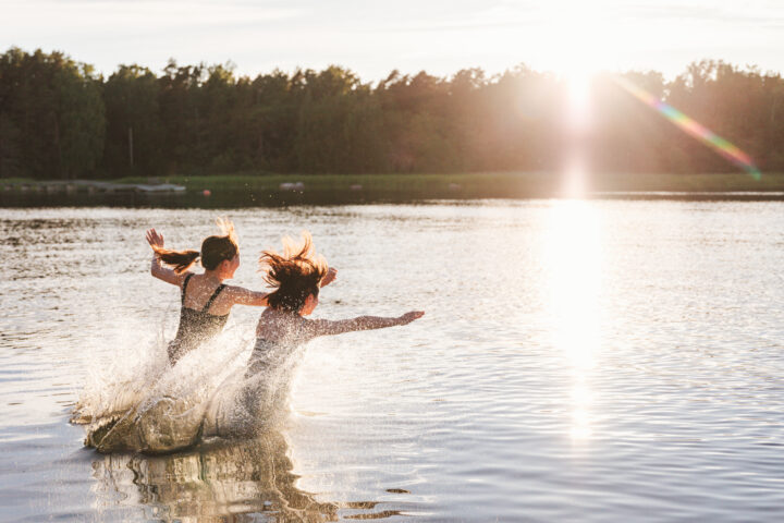 Two girls joyfully splash into a lake at sunset, with sun flare.