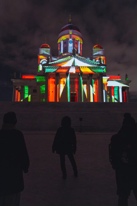 Helsinki Cathedral illuminated with vibrant green, red, and white lights at night, with silhouetted figures in the foreground.