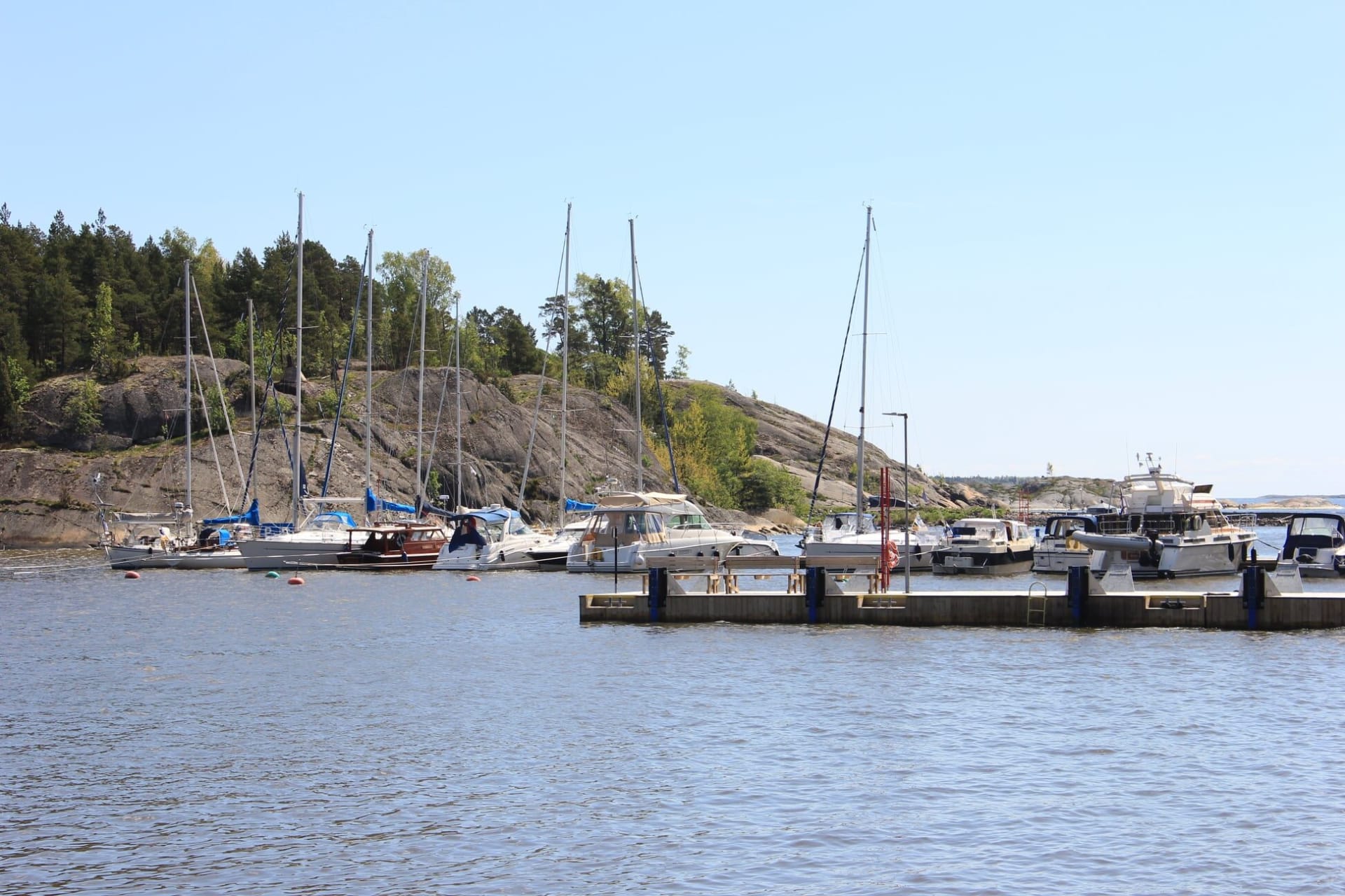 A sunny marina with several moored motorboats and yachts, with people standing on the dock. A gravel path runs along the waterfront, with visitors walking toward the harbor buildings surrounded by trees.