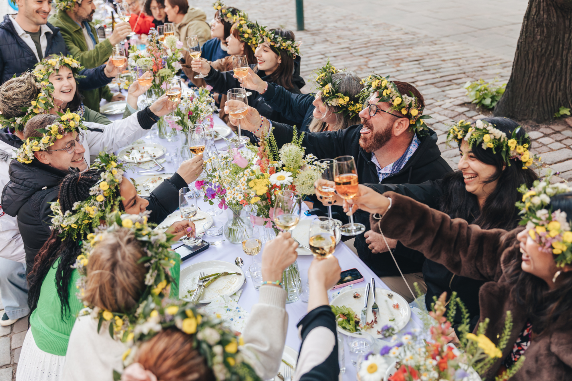 People in flower crowns toast with drinks at a long outdoor table.