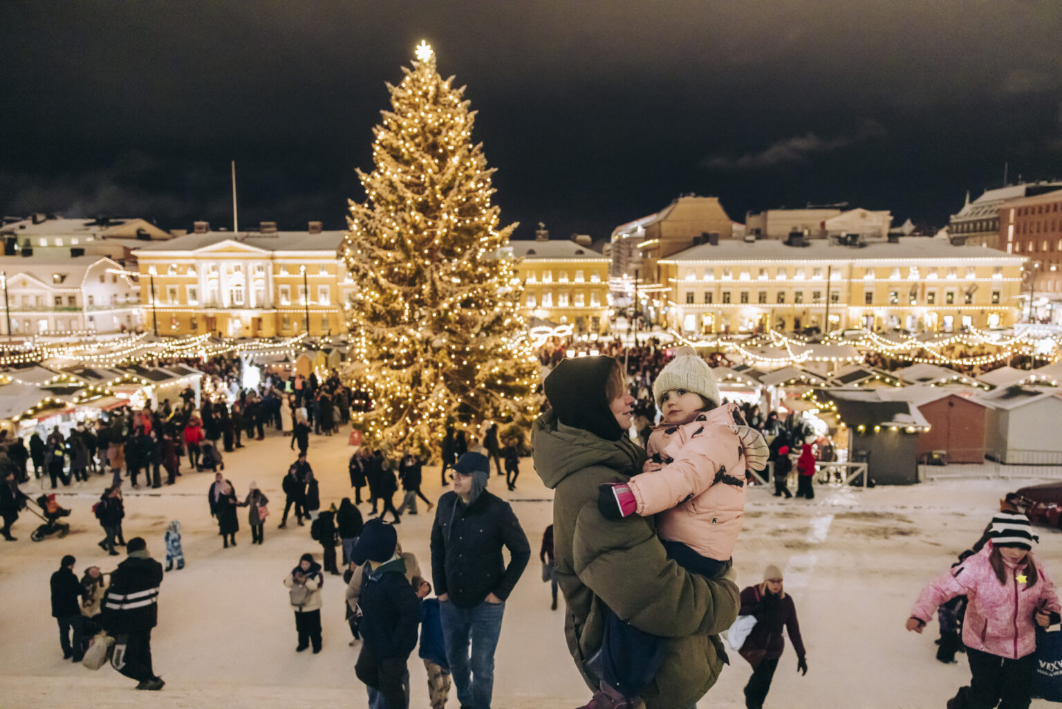 Family enjoys a festive Christmas market with a large decorated tree in the background.