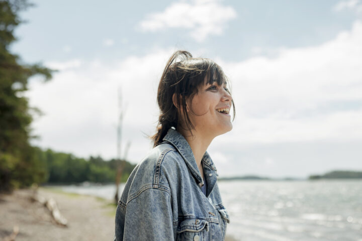 Young woman in denim jacket laughing by a lake on a sunny day