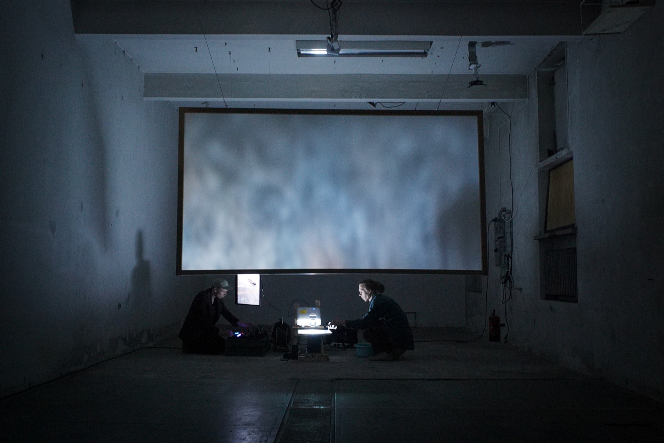 Näyttelyn tiedotuskuva, jossa kaksi ihmistä hämärässä tilassa.