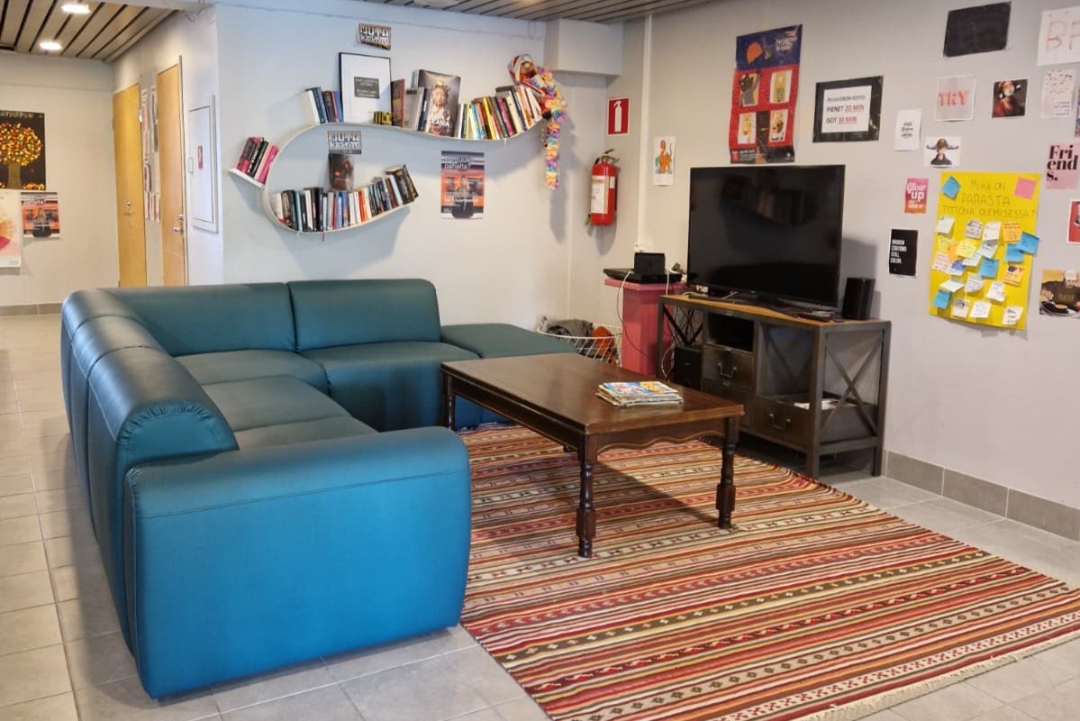 Kuvassa turkoosi kulmasohva ja kuvan takareunassa on kirjahylly täynnä kirjoja. Oikealla seinustalla värikkäitä julisteita sekä musta televisio.