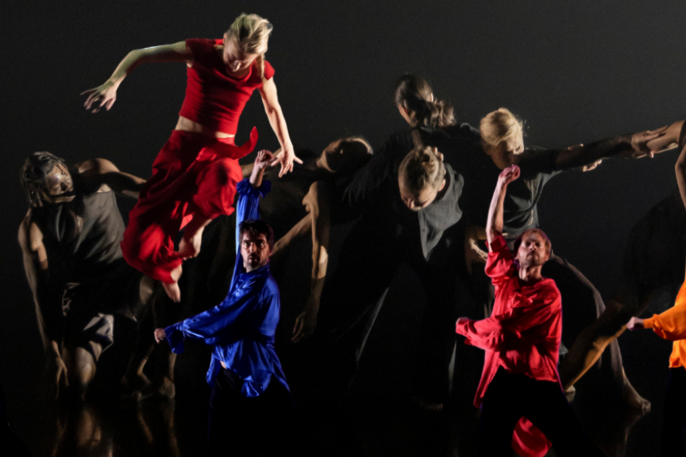 Tanssikohtauksia kolmesta eri esityksestä, etualalla kaksi miestä värikkäsiiin vaatteisiin puettuna käsi ylhäällä, taustalla mustiin pukeutuneita nykytanssijoita ja vasemmassa reunassa punaisiin pukeutunut hyppäävä naistanssija.