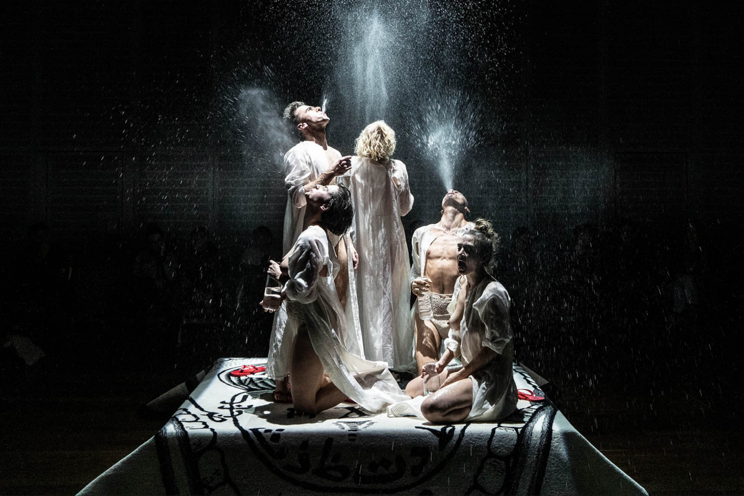 Kolme naista ja kaksi miestä sylkee vettä ylöspäin korokkeen päällä.