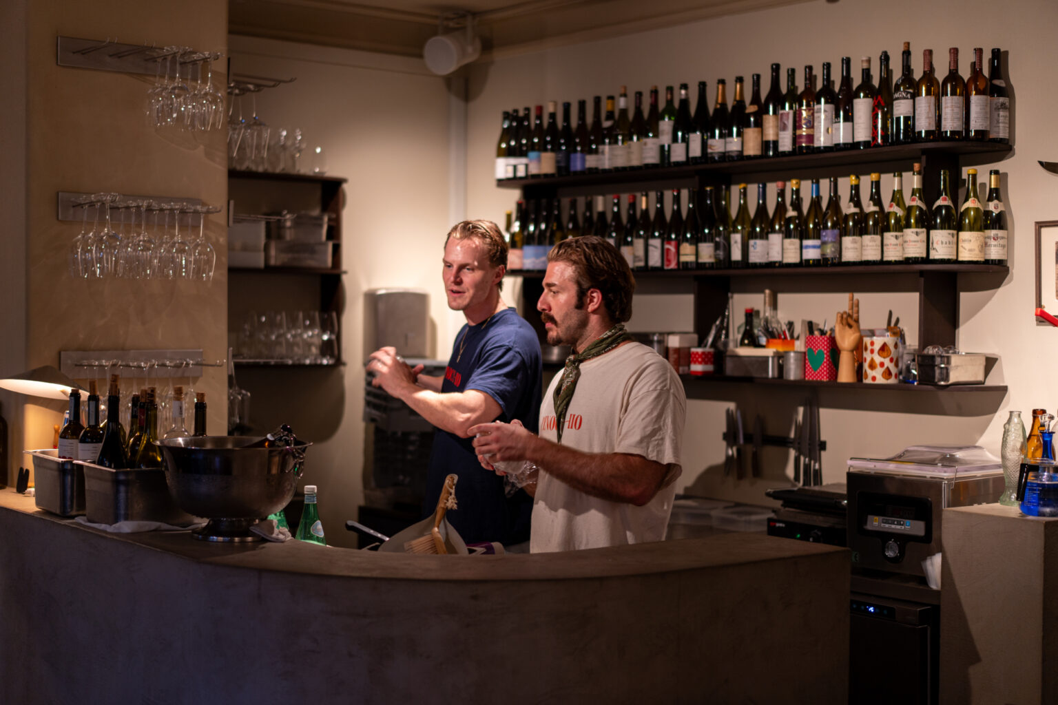 Two men behind a bar, one wearing a blue t-shirt, the other a white t-shirt with text.