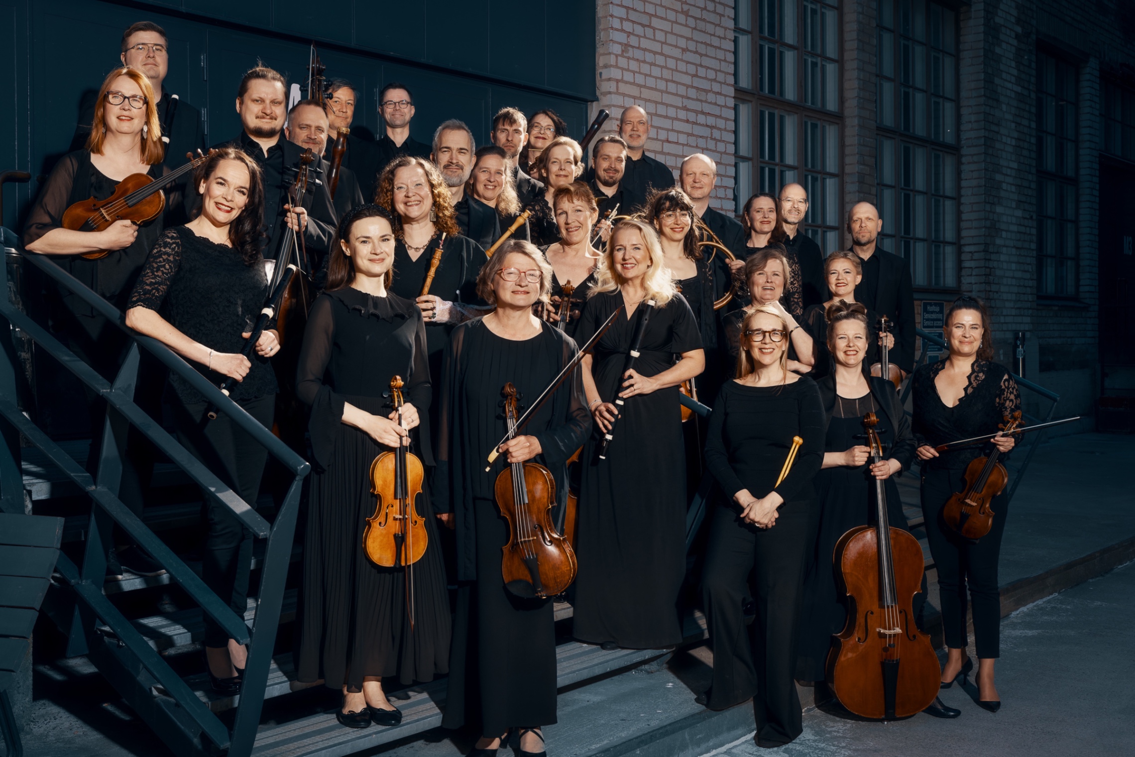 Noin 30 Suomalaisen barokkiorkesterin muusikkoa mustissa esiintymisvaatteissaan ja soittimineen poseeraa kuvaajalle