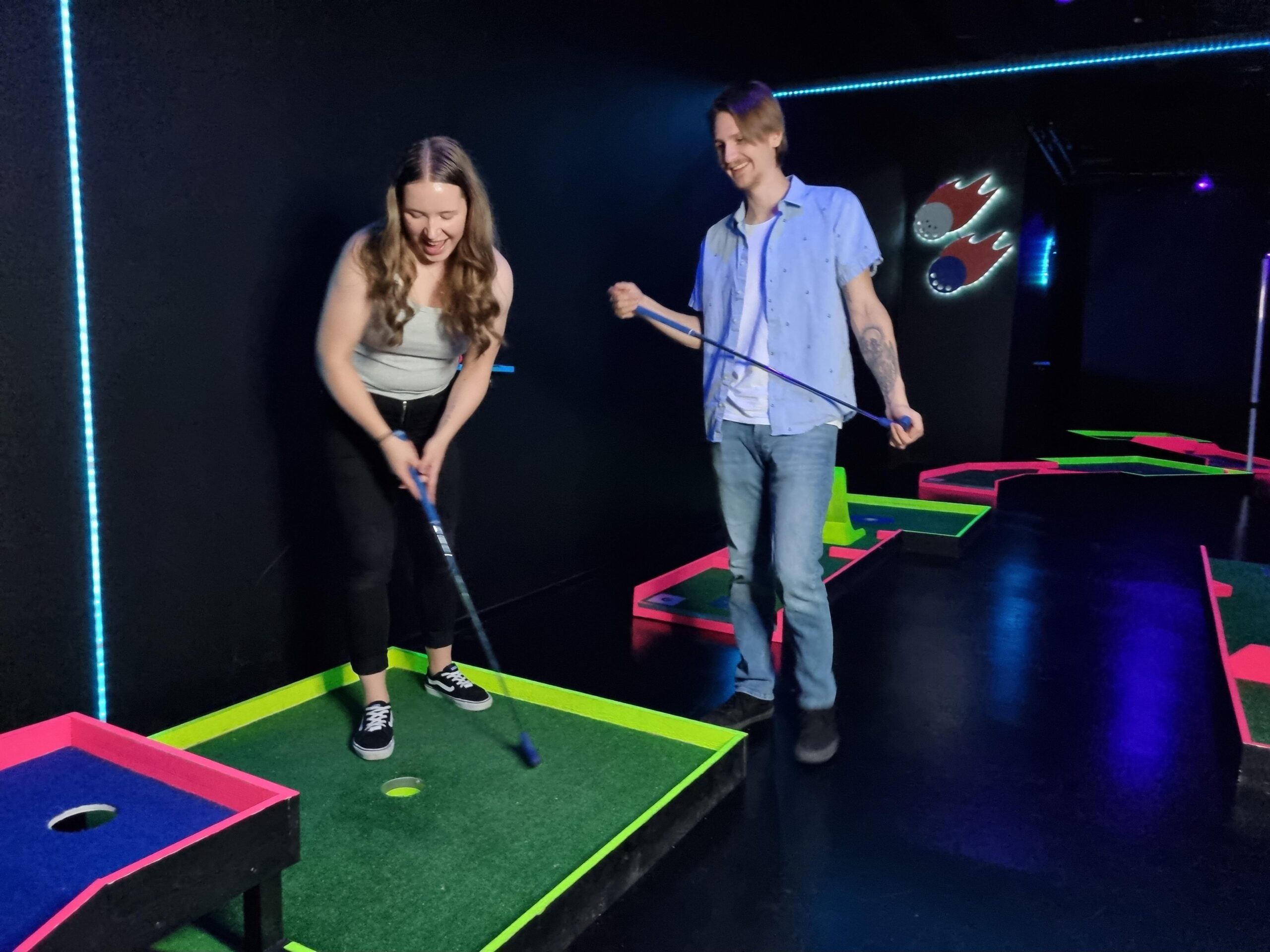 Två vuxna spelar minigolf på Hohtogolf REDIs neonbelysta bana och skrattar tillsammans medan de puttar bollen i hålet.