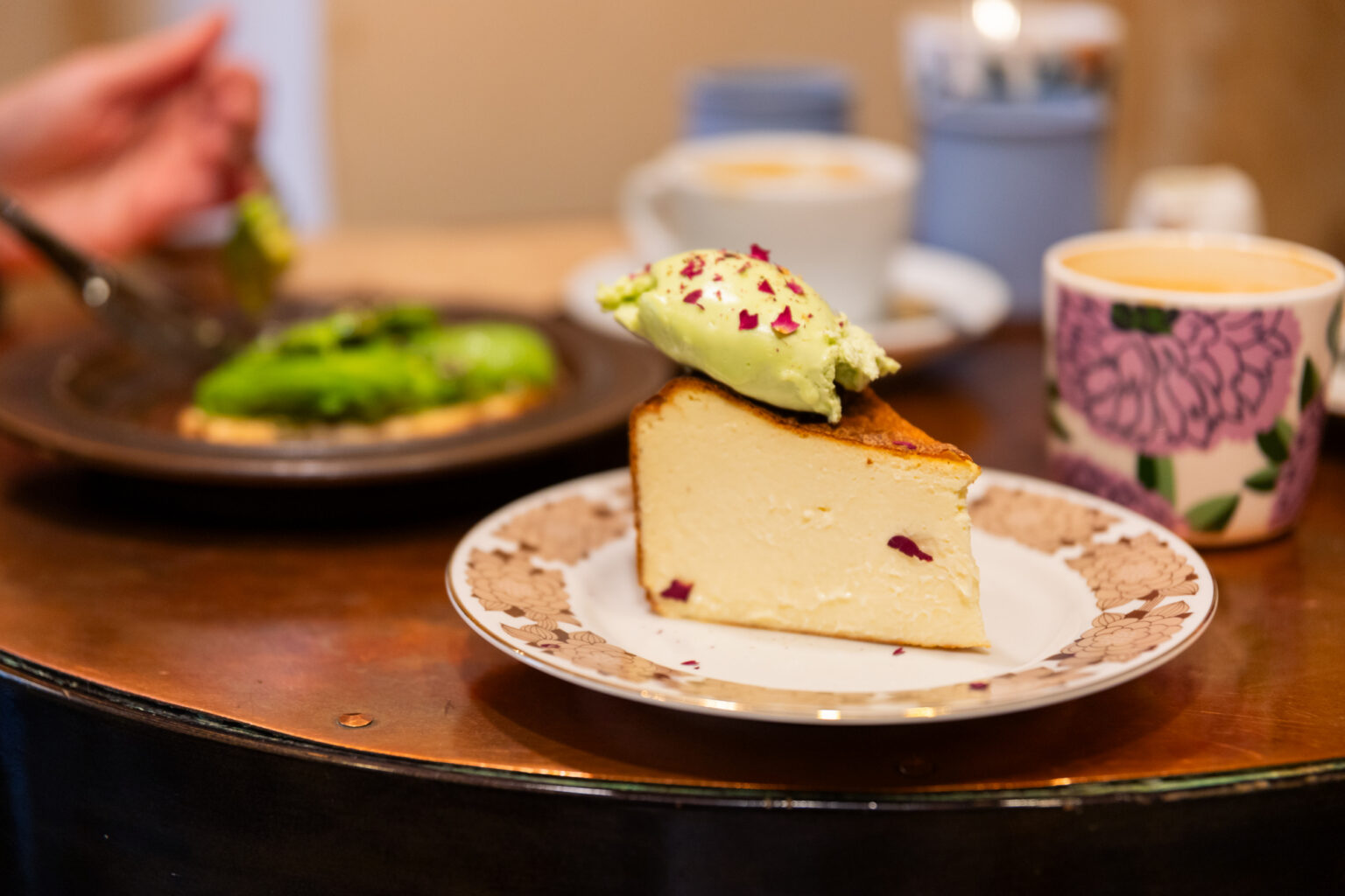 Slice of cheesecake with green topping on a plate, with coffee and pastry in background