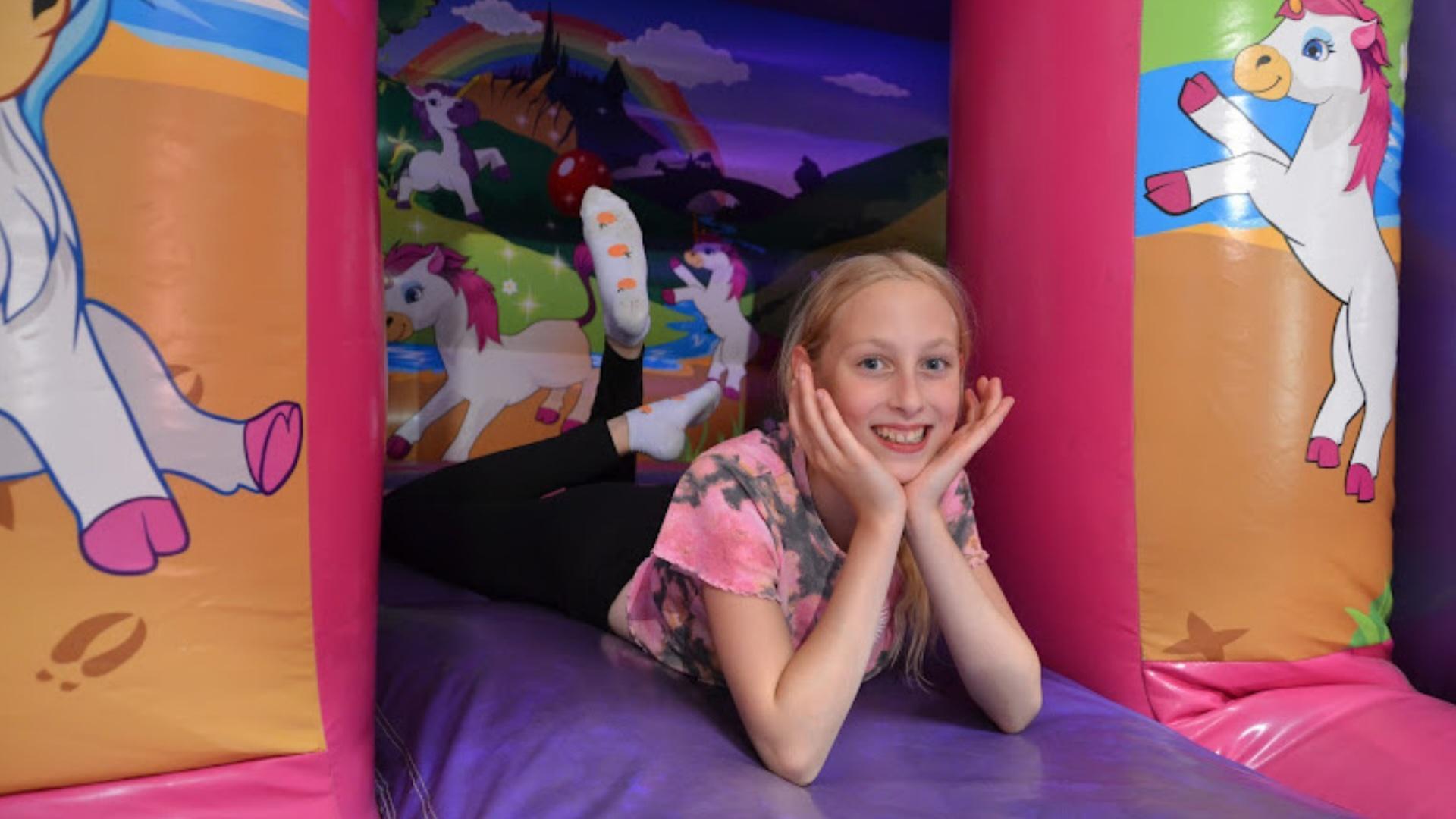 Hymyilevä lapsi makaa pomppulinnassa värikkäiden yksisarviskuvitusten ympäröimänä ja katsoo kameraan iloisena.