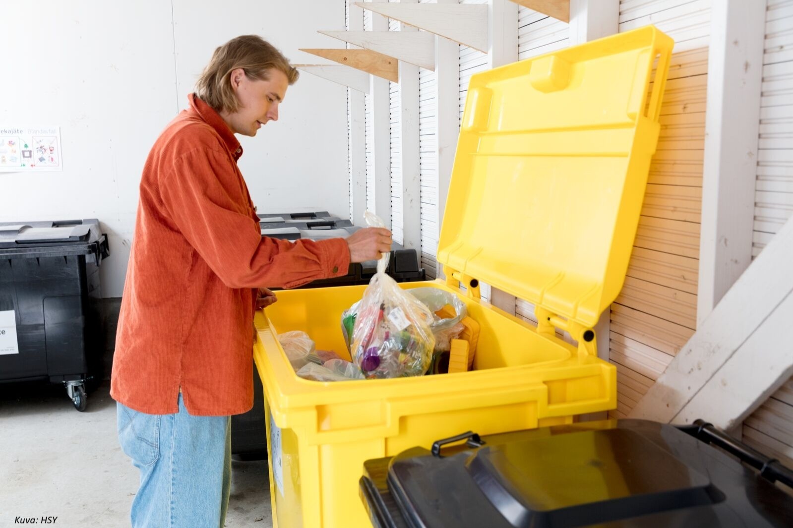 Henkilö pudottaa muovipussillista muovijätettä keltaiseen muovinkeräysastiaan.