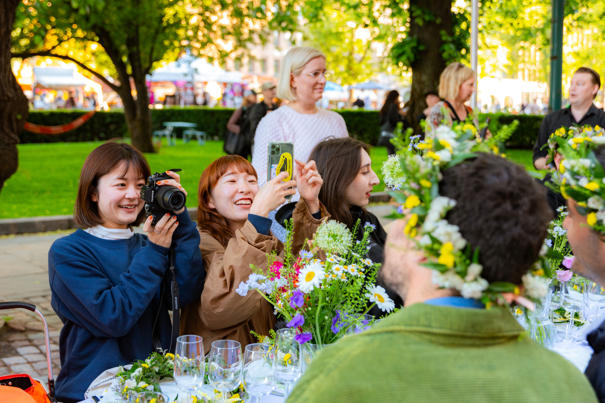 People at an outdoor table with flowers, one woman taking a photo