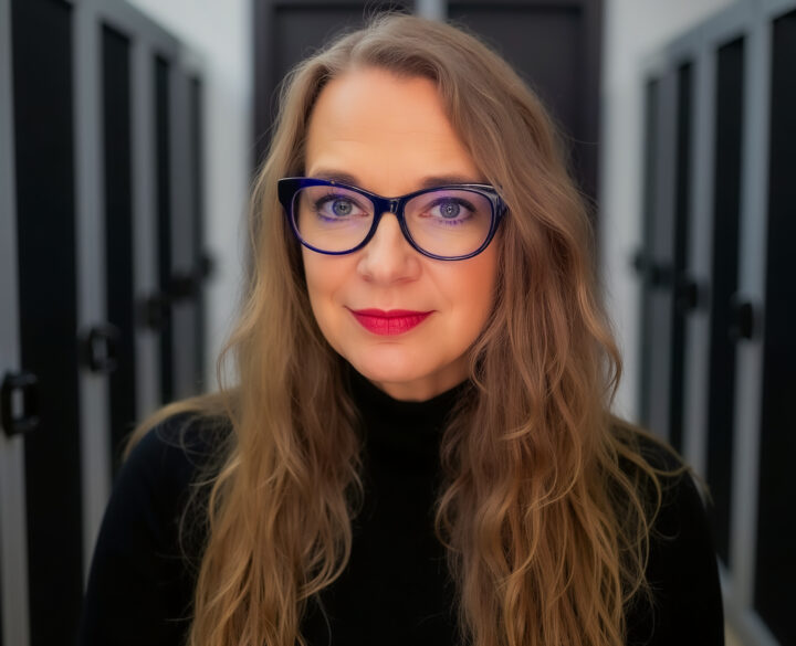 Woman with long blonde hair and blue glasses smiles in front of lockers