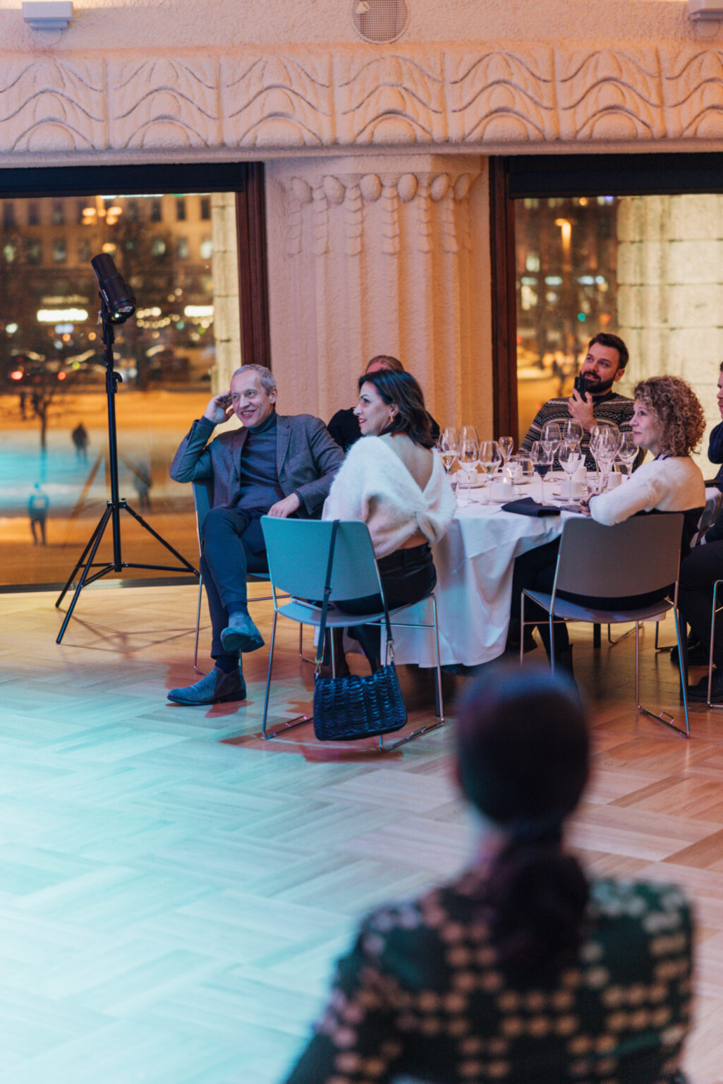 People dining and conversing at a formal event with city lights visible through large windows.