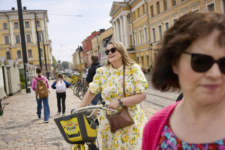 Woman in lemon dress rides yellow bike past cobblestone street and historic buildings in Helsinki