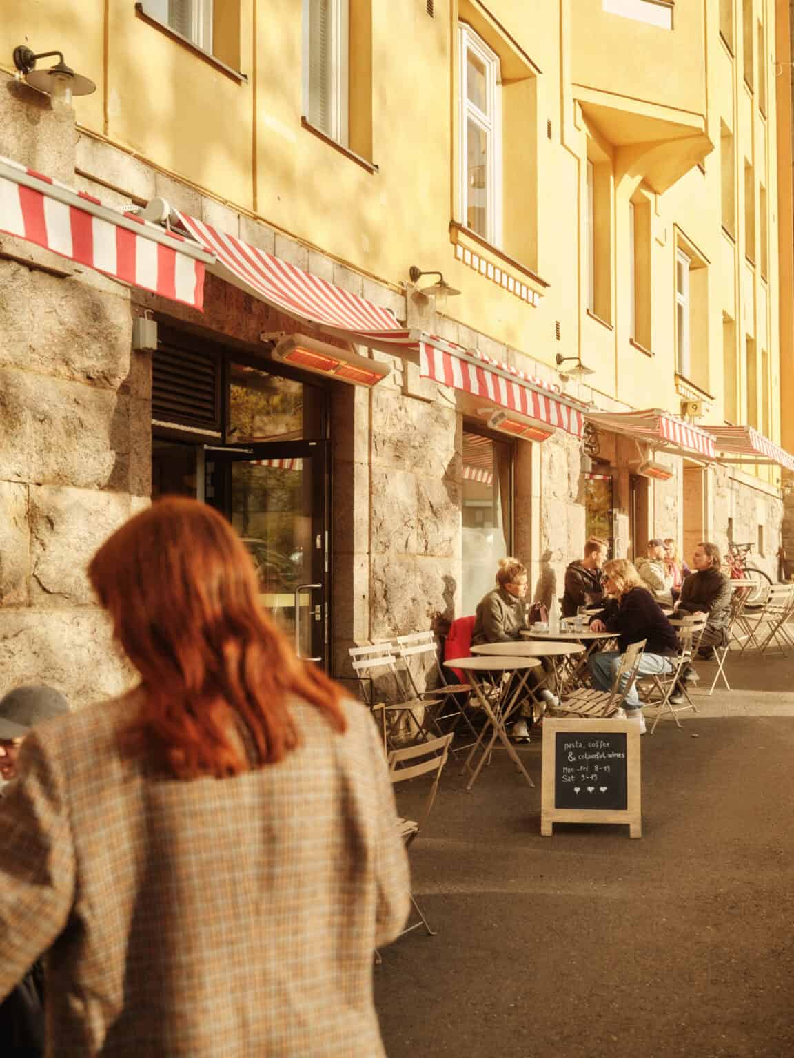 Outdoor cafe with people dining under red and white awnings; menu board lists pasta, coffee, wine.