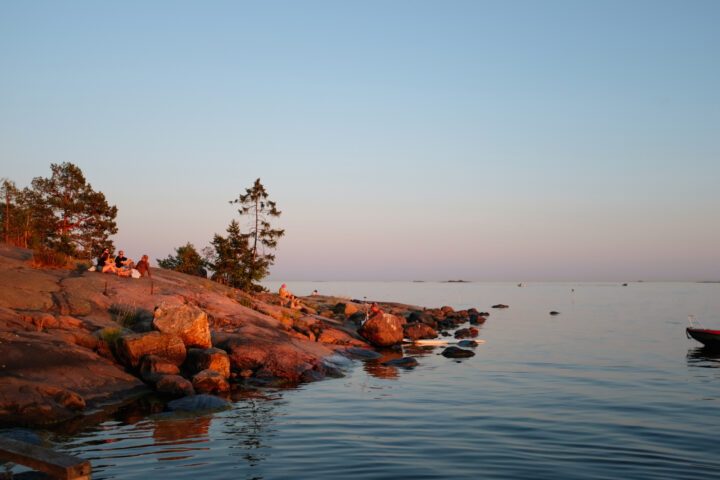People relax on a rocky shore at sunset, overlooking a calm sea with boats.