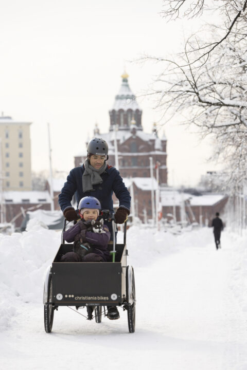Father and child in a Christiania cargo bike in the snow, with a building in the background.