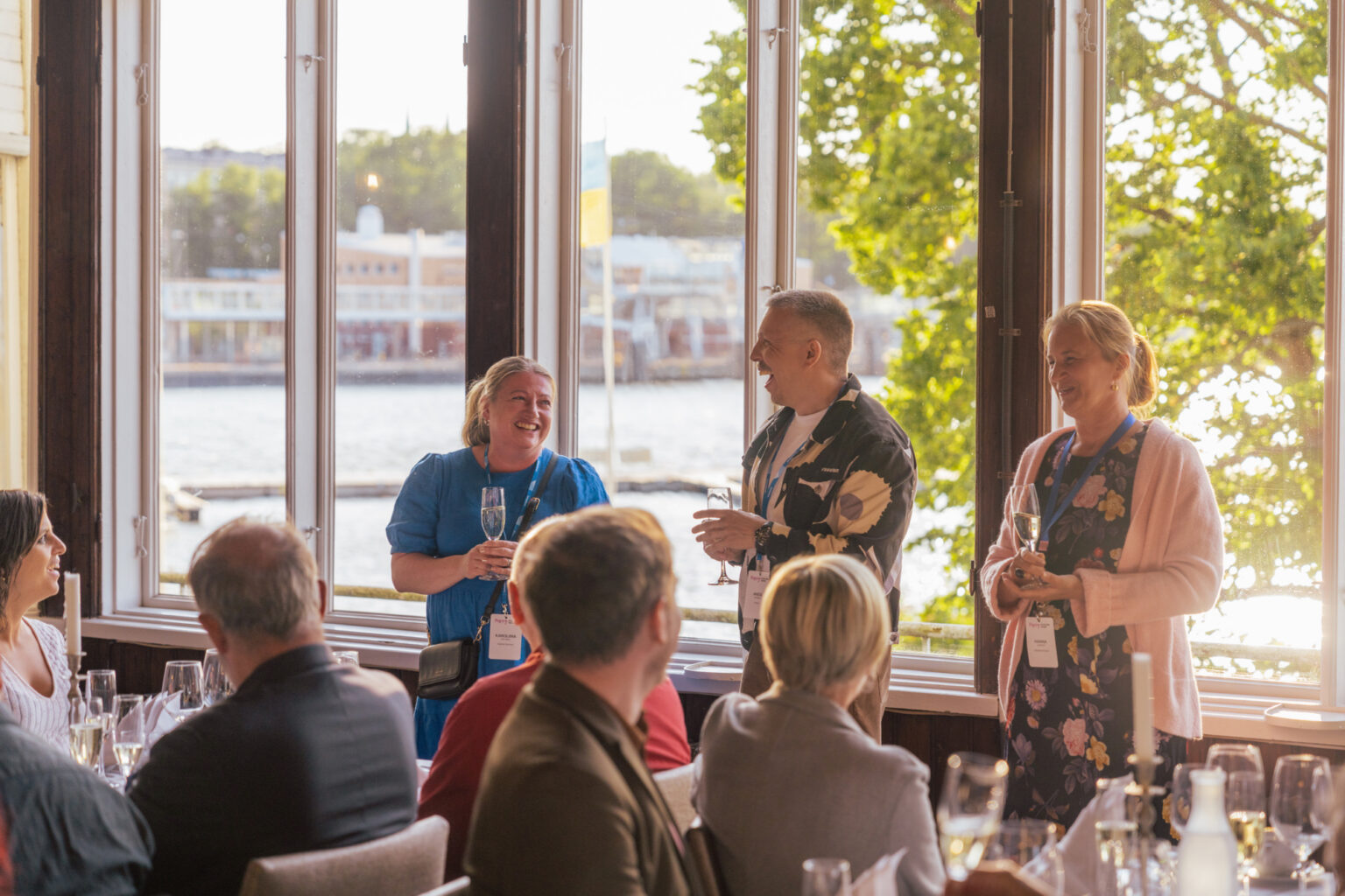 People mingling and enjoying drinks at a waterfront event with large windows