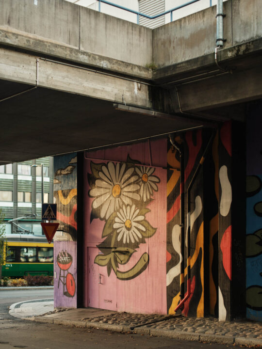 Graffititaideteos vaaleanpunaisella seinällä betonisen ylikulkusillan alla, jonka ohi kulkee raitiovaunu.
