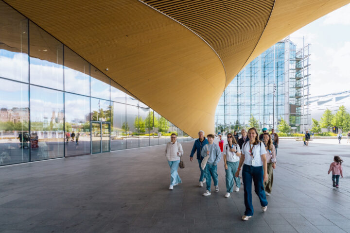 Group of tourists walking past modern architecture with curved wooden ceiling
