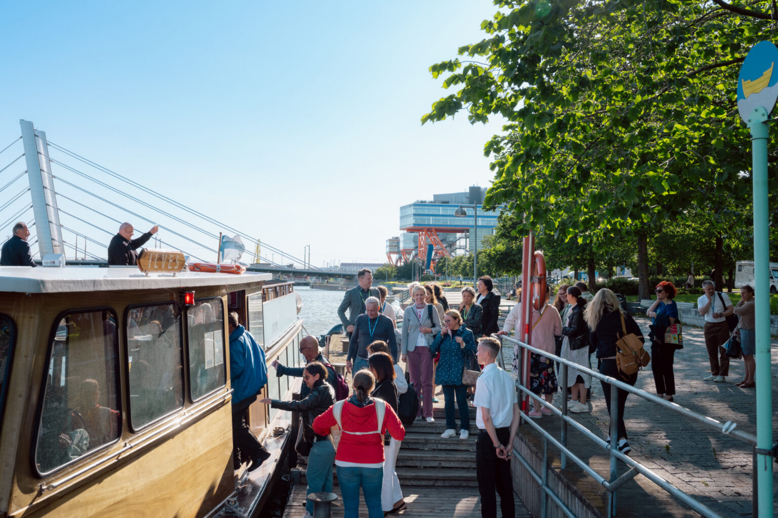Tourists board a boat tour in a sunny European city, with modern architecture in the background.