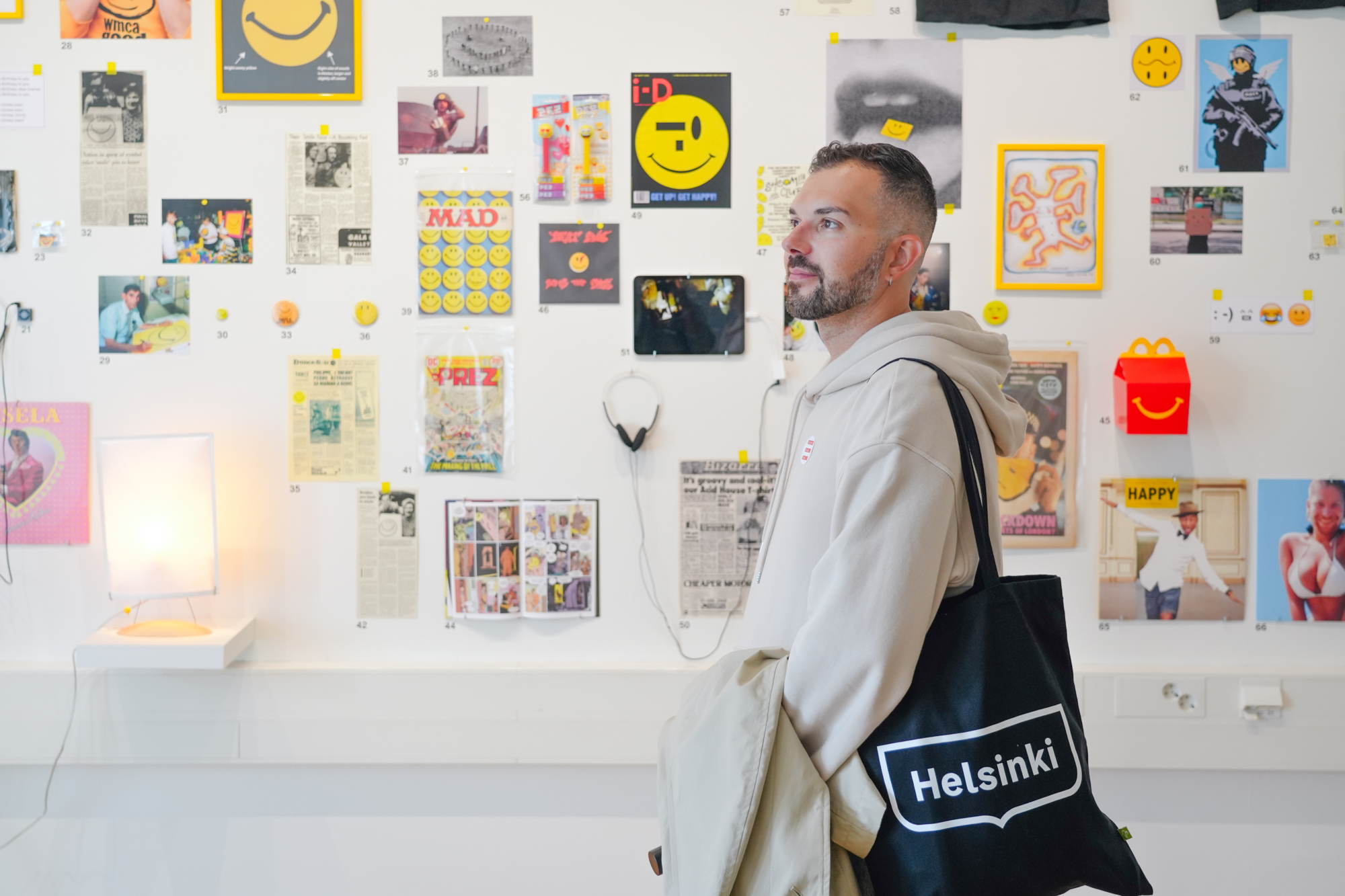 Mies, jolla on Helsinki-laukku, seisoo taideteosten ja hymynaamojen koristamien julisteiden edessä.