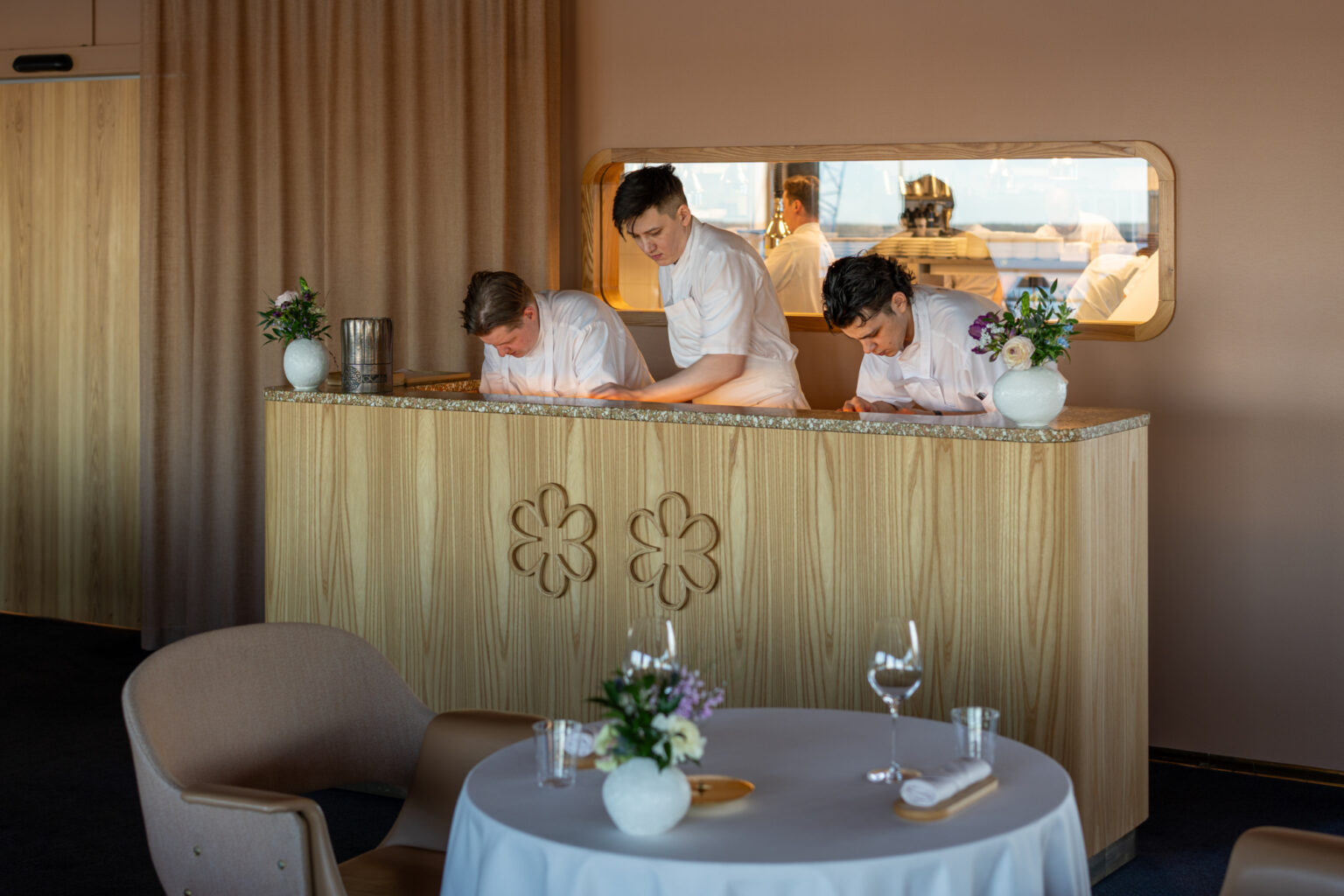 Chefs in white uniforms lean over a counter in a modern restaurant.