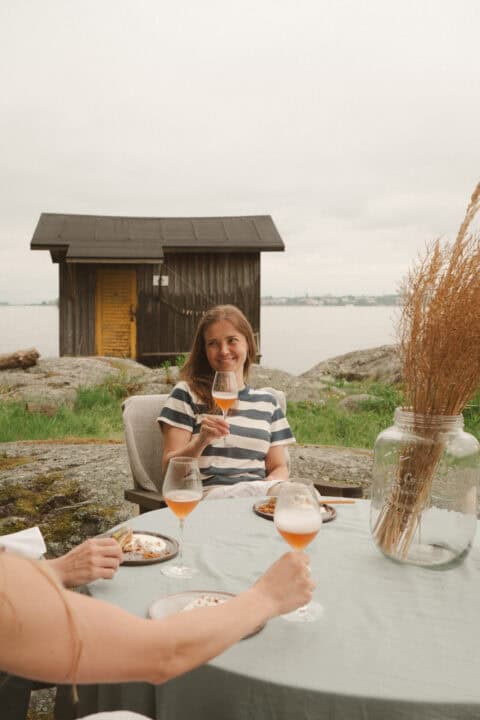 Woman smiling and holding a glass of beer at an outdoor table near a wooden shed.